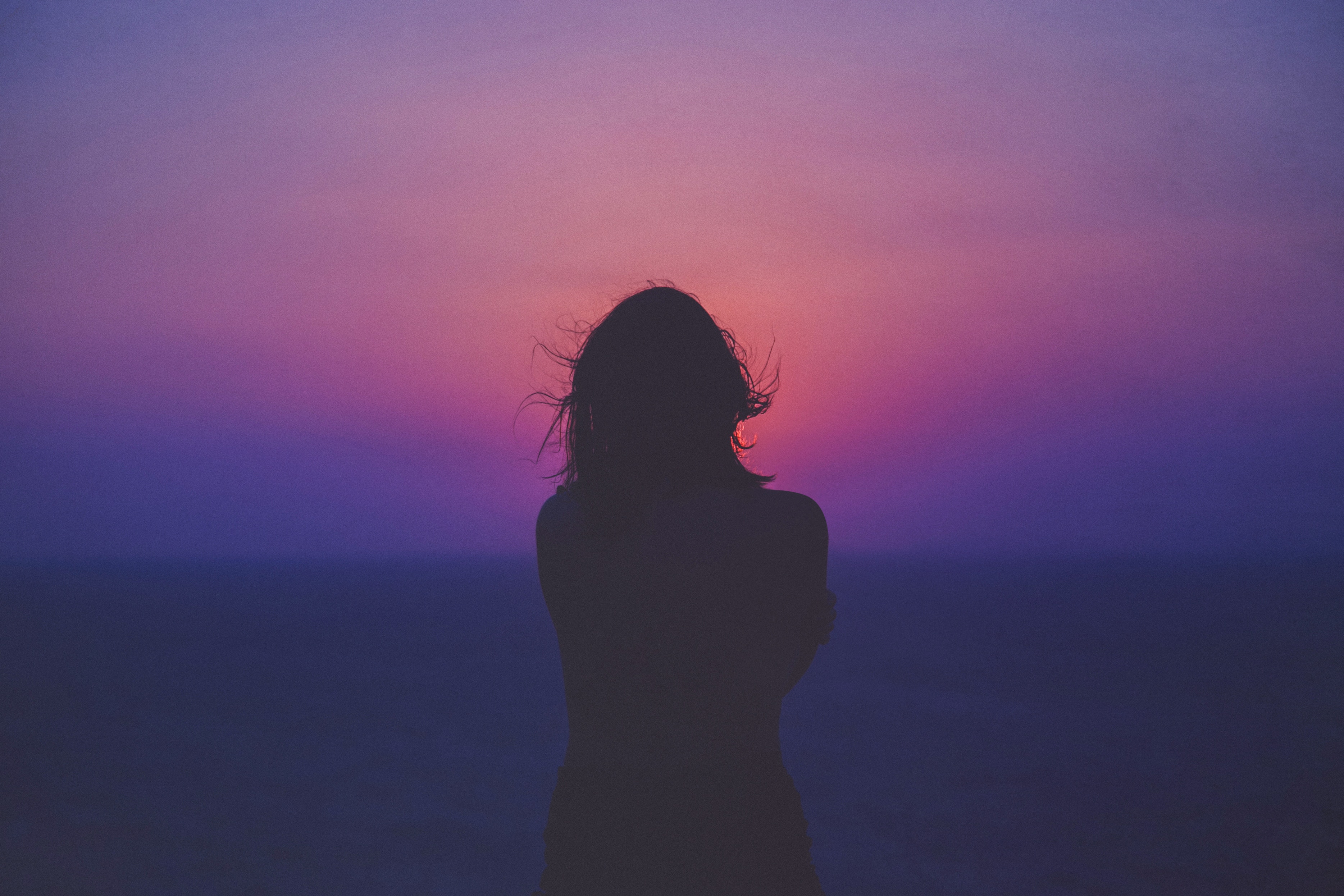 silhouette of woman's back  standing windy facing a hazy sunset mostly blueish pinkish colors, hazy