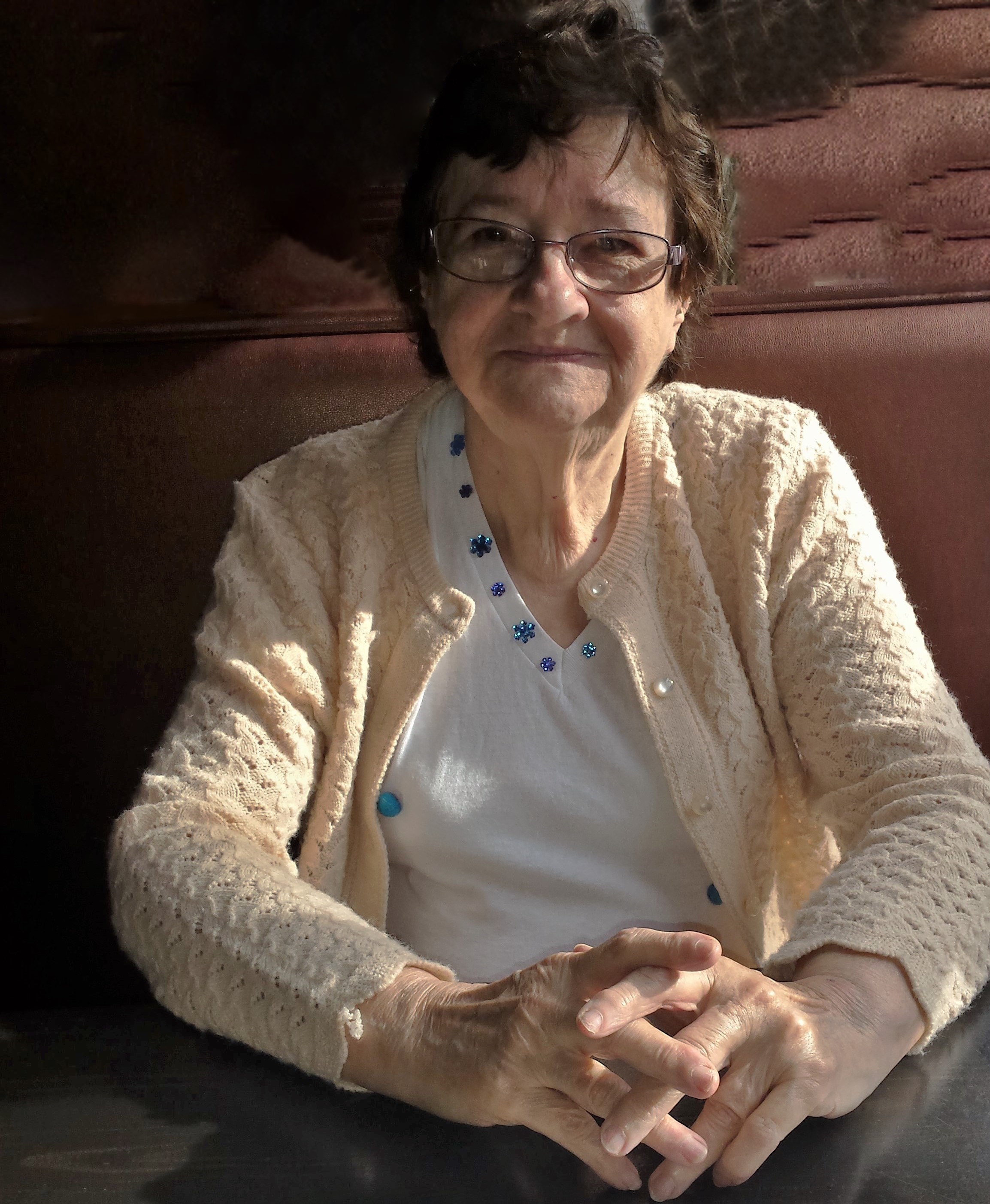 photo of Janet Hurlow, facing camera, tan sweater, white blouse, smile, slightly tinged glasses black hair, sitting at a table with hands folded brown booth in diner