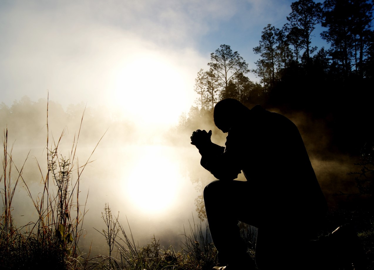 silhouette of man kneeling and praying in front of lake with woods surrounding it sunrise mostly white with some fog