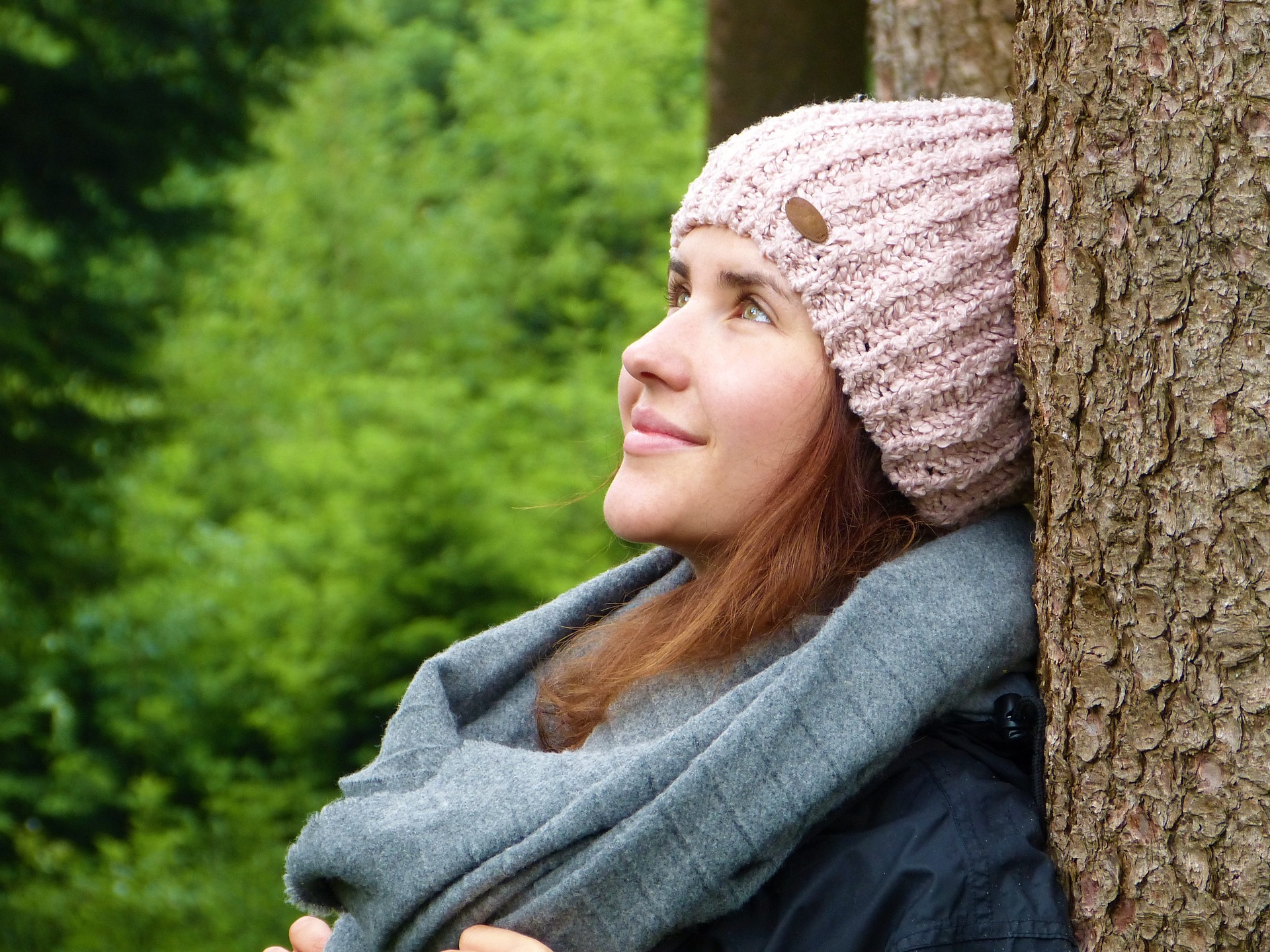 young woman leaning against tree looking up trees in background, pink sweater hat, smiling,