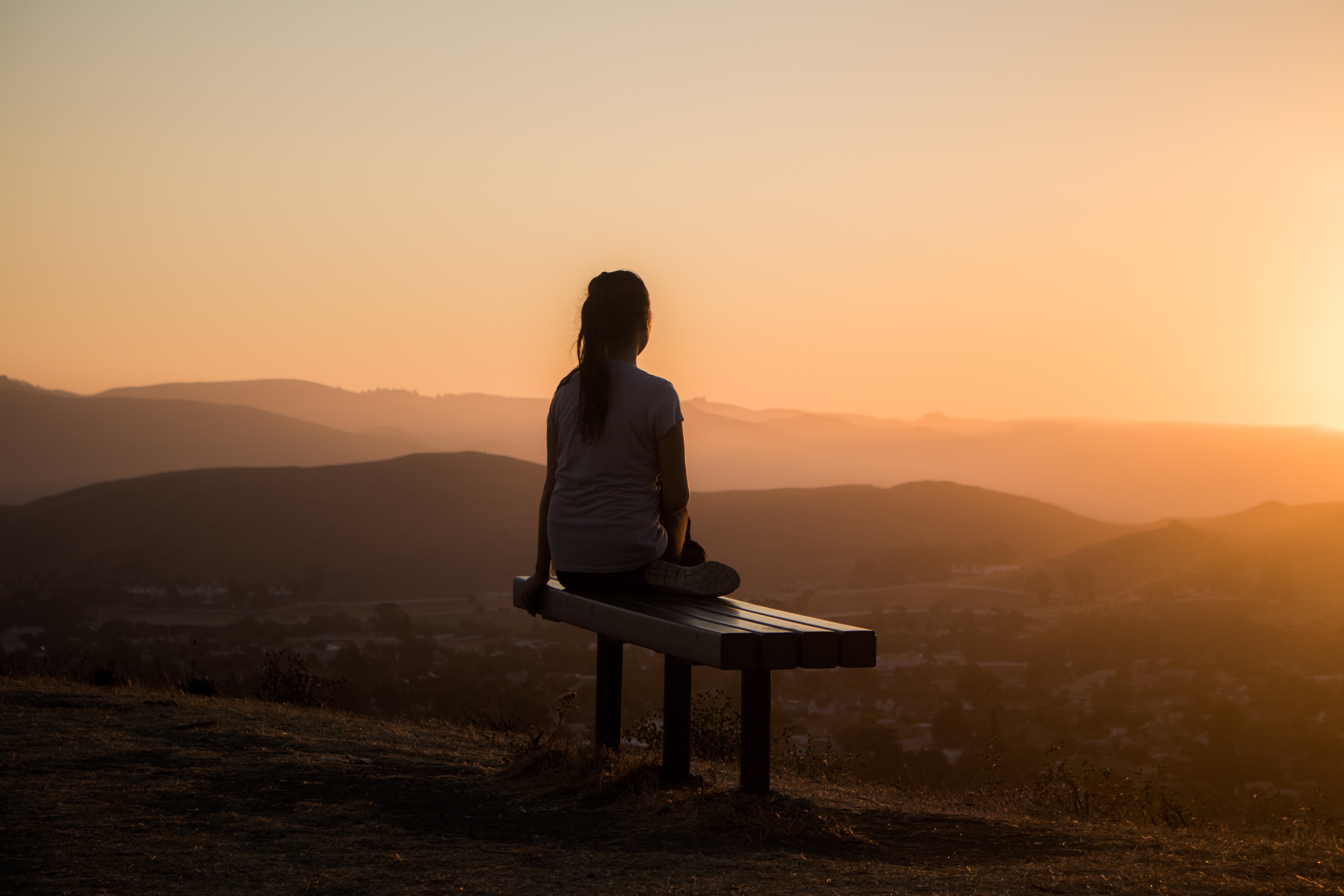 young woman sitting on bench facing the sunrise over the mountains, yellowish