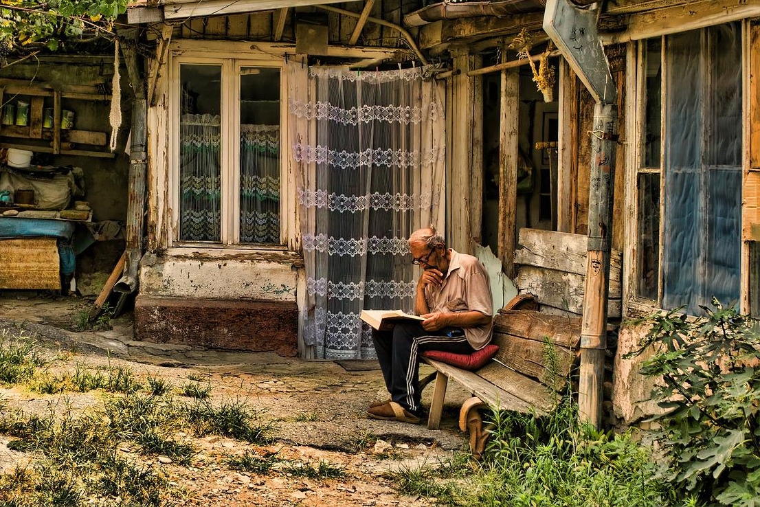 old man sitting on bench beside old house reading wood shack weeds and rocks